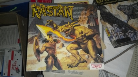 A boxed copy of the game Rastan, which shows a man with fire coming off his sword to strike a lizard-thing